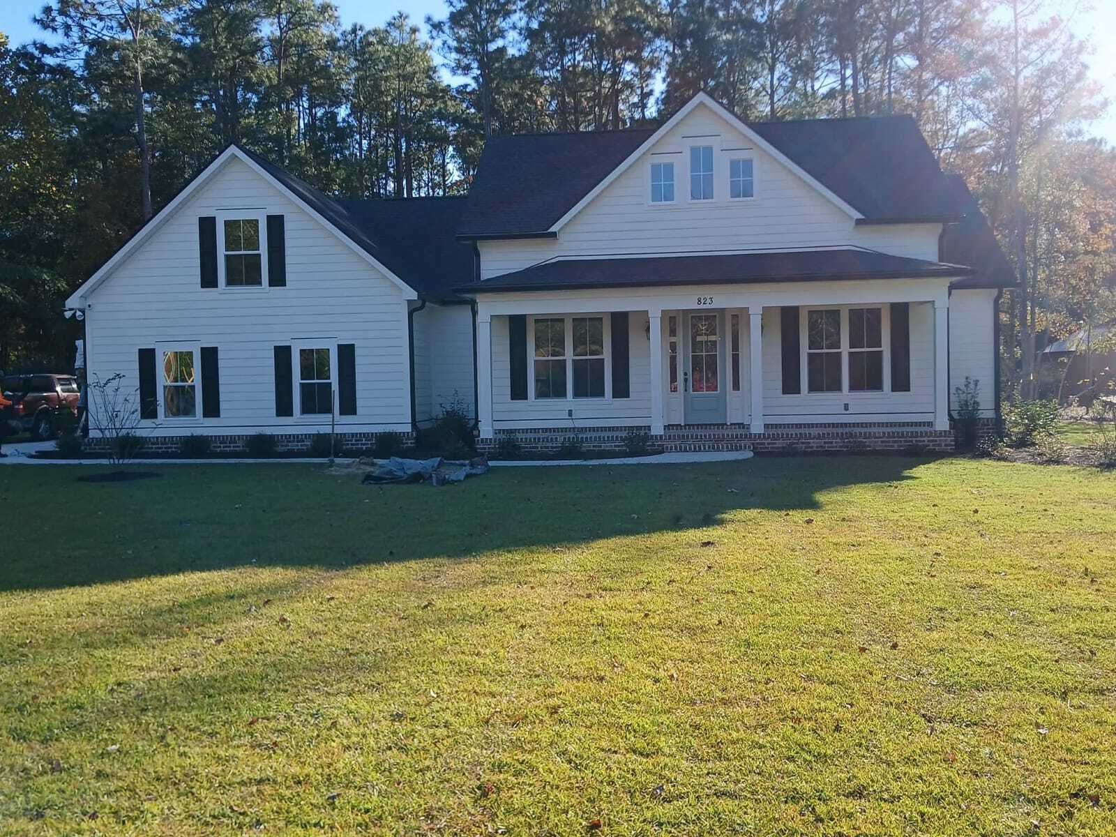 A white, two-story house with dark shutters sits in a sunny yard surrounded by tall trees. The home has a welcoming porch and vibrant green lawn.