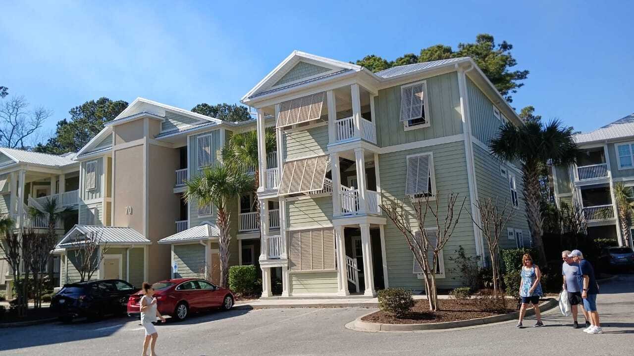 Three-story pastel green building with white trim and balconies, surrounded by sparse landscaping and palm trees. People walk in bright, sunny weather.