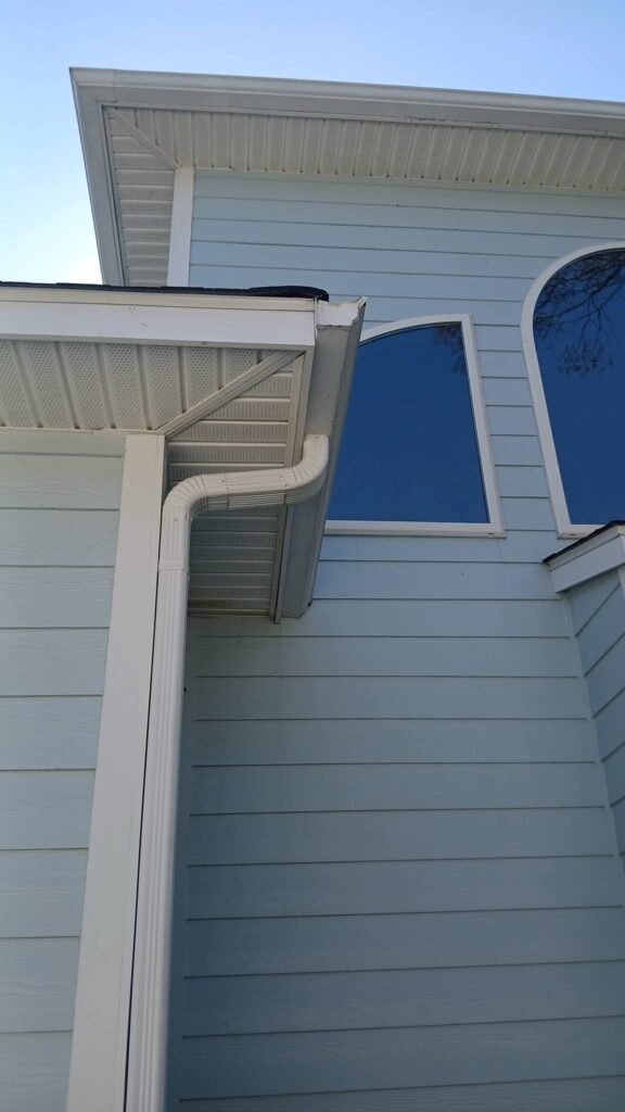 The image shows a corner of a light blue house with white trim, featuring a downspout and two tall, arched windows reflecting a clear blue sky.