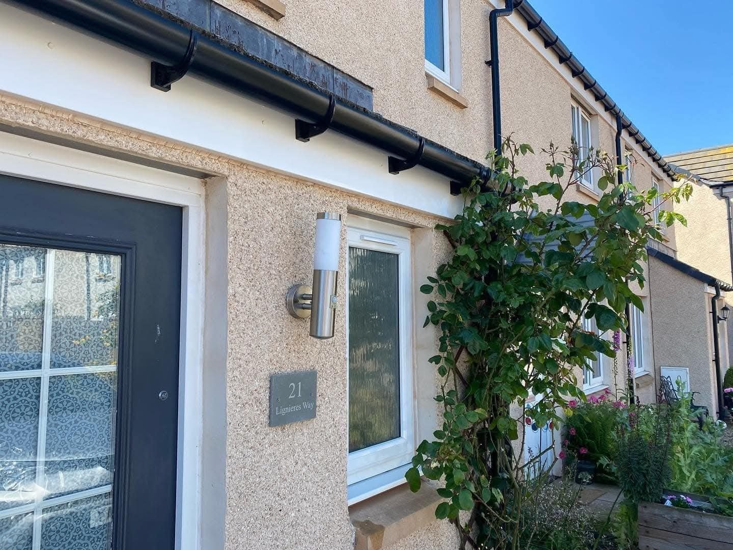 House exterior in sunlight with beige walls, black roof trim, and a window next to a door. A number 21 plaque and a tall leafy plant are present.