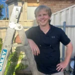 Smiling person in a black shirt leans on a ladder in a backyard. Sunlight casts shadows on the light-colored fence and brick house, creating a warm atmosphere.