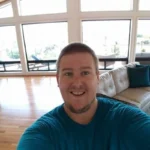 A smiling person wearing a blue shirt takes a selfie in a bright, spacious living room with hardwood floors, large windows, and a white sofa.