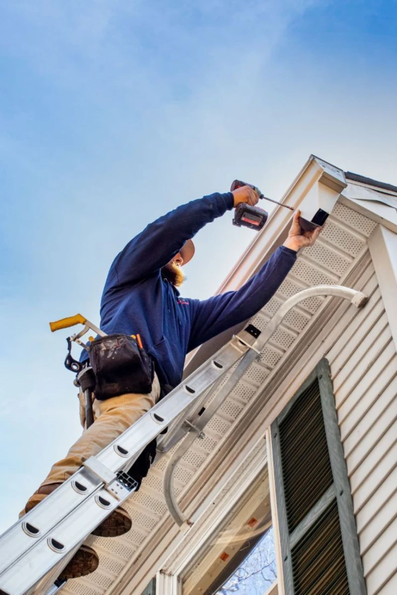 A worker wearing a blue jacket and tool belt stands on a ladder, using a power drill on a house's exterior gutter. The sky is clear and blue.