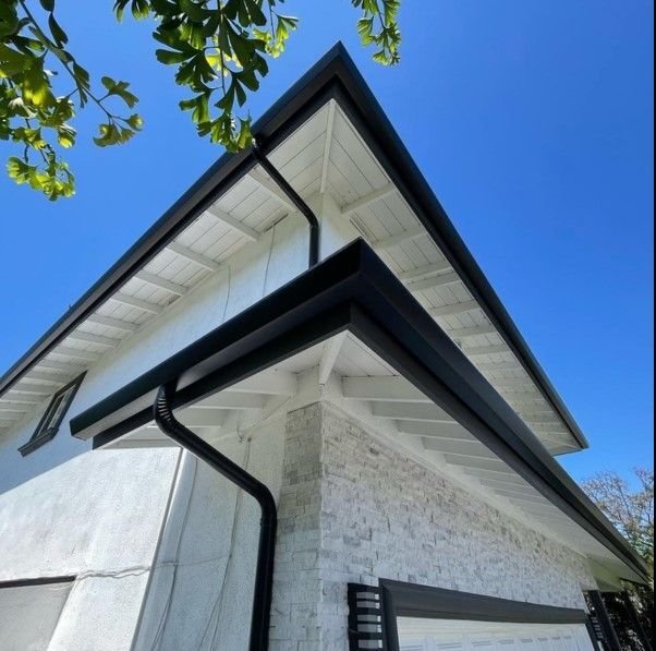 Close-up of a modern house corner under a clear blue sky. Features a black gutter, white brick wall, and green leaves in the top left. Clean and fresh tone.