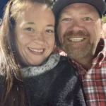 Smiling man and woman close together, outdoors at night. She wears a dark sweater, he a red plaid shirt and cap. Warm, happy atmosphere.