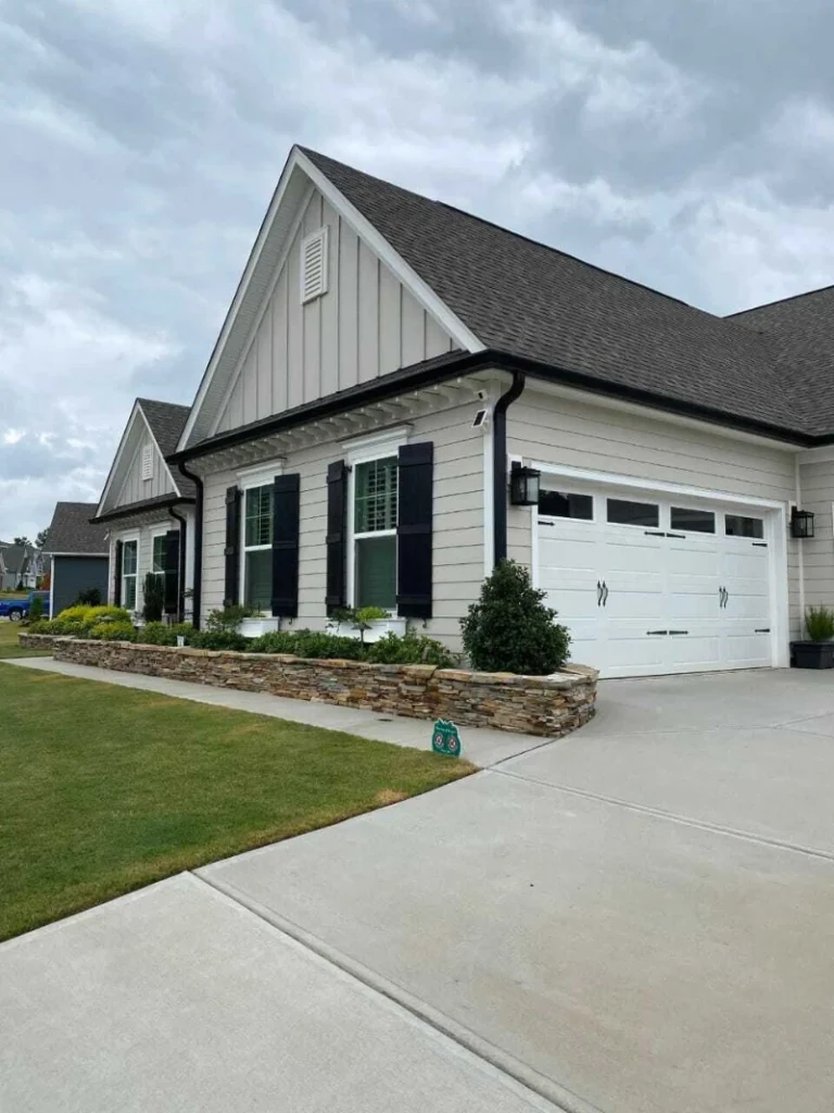 Single-story house with gray siding, black shutters, and a white garage. Stone-bordered landscaping and cloudy sky create a calm suburban feel.