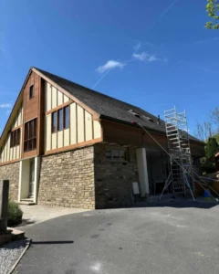 A two-story house with a stone and wood exterior is shown under a clear blue sky. A scaffolding stands against the side, suggesting ongoing construction.