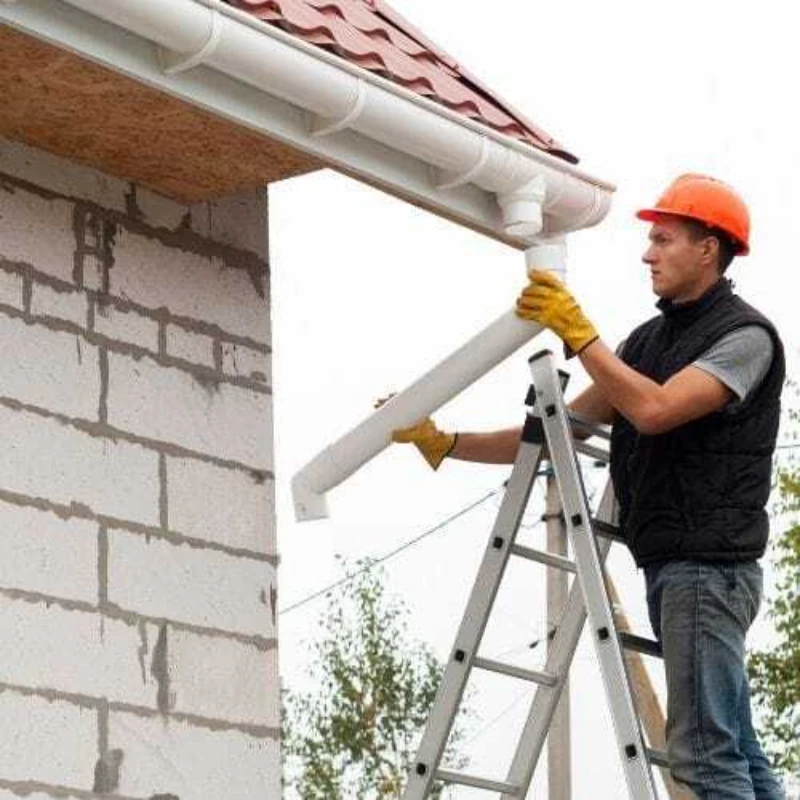 A construction worker in orange helmet and yellow gloves stands on a ladder, installing a white gutter on a brick house with a red tiled roof.