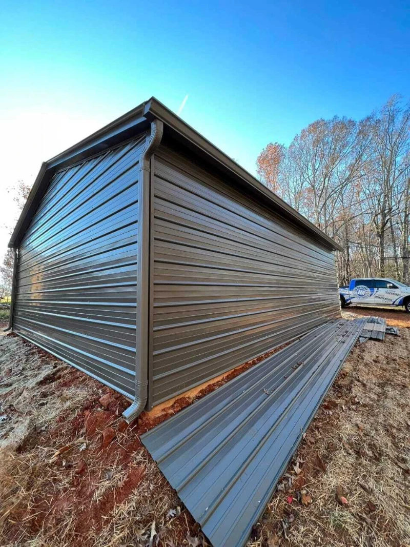 A large, newly installed brown metal shed stands on a grassy, slightly sloped terrain. Piles of unused metal panels lie beside it. Trees and a utility vehicle are visible in the background under a clear blue sky.