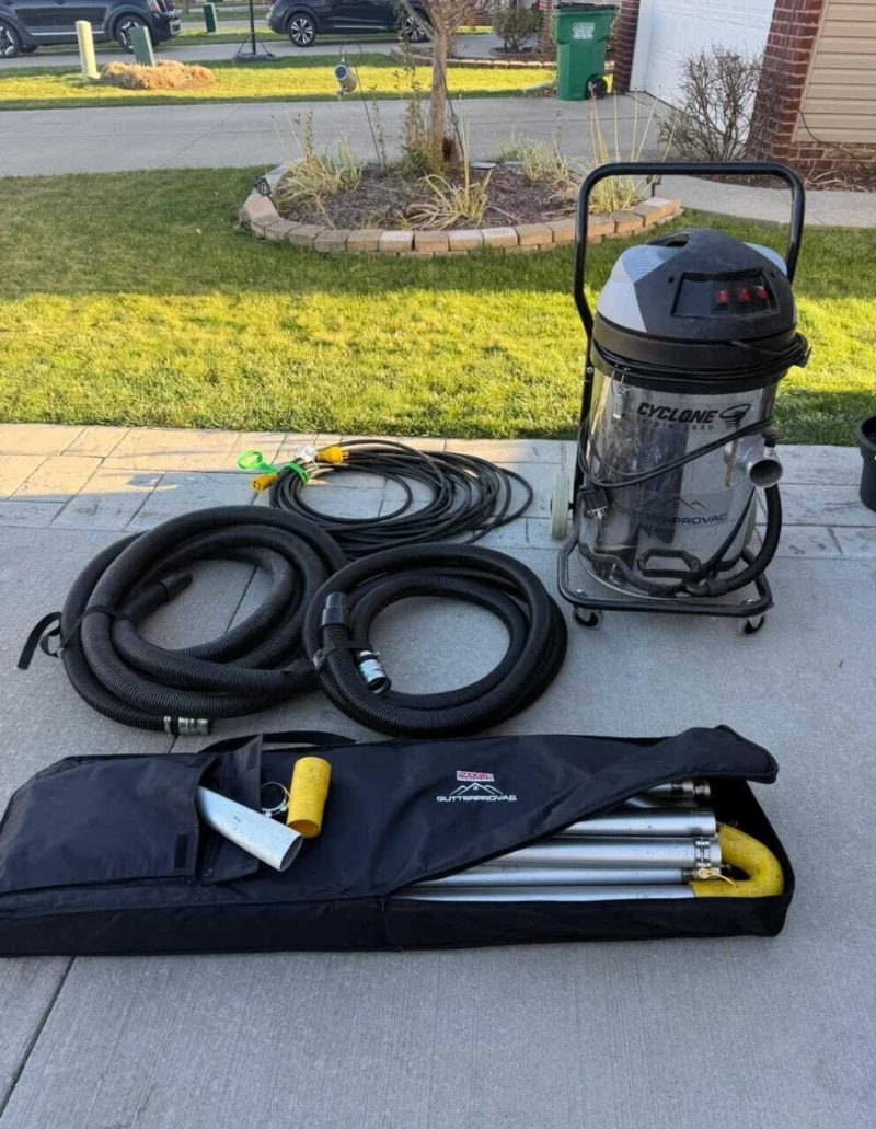 A heavy-duty vacuum cleaner is on a driveway, surrounded by long hoses and a black bag containing metal poles. Grass and garden beds in the background.