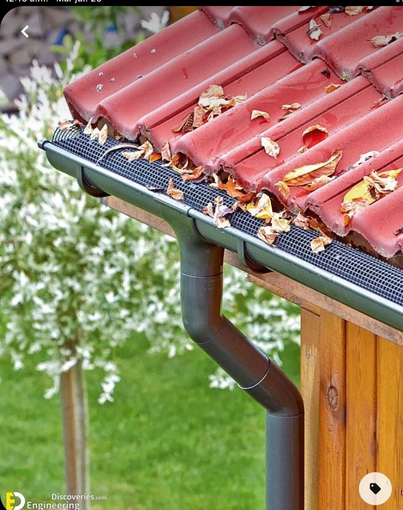 Red roof tiles with scattered autumn leaves on a roof gutter equipped with a mesh guard. A black downspout is seen against a blurred green garden.