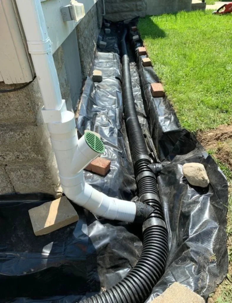 White PVC pipe connects to black drainage pipe along a wall, surrounded by black tarp and red bricks. Adjacent is a patch of green grass.