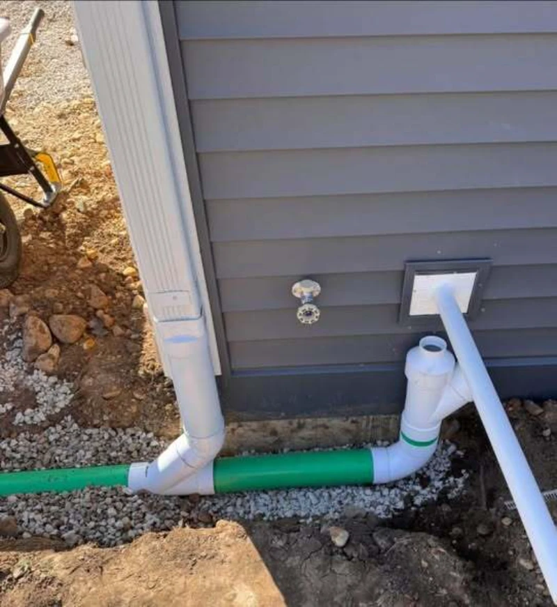 Exterior view of a building corner with gray siding, featuring white and green drainage pipes. A wheelbarrow with rocks is visible nearby on the gravel.