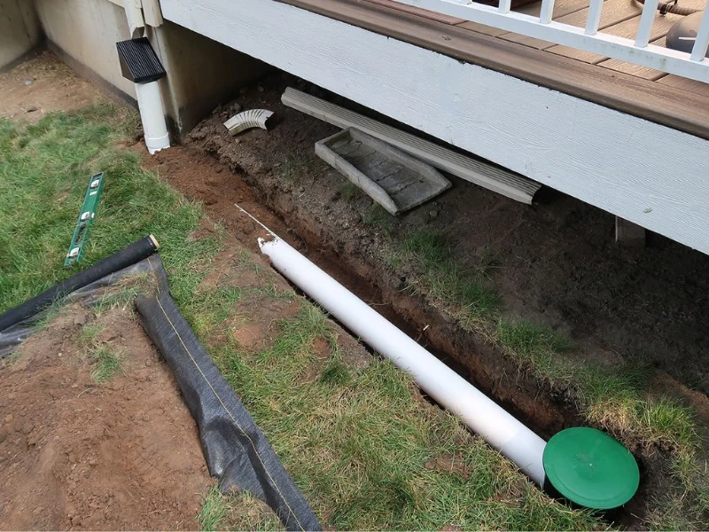 A yard trench with a partially buried white PVC pipe, a green cap, and nearby tools, bordered by grass and a wooden deck, conveying home renovation.