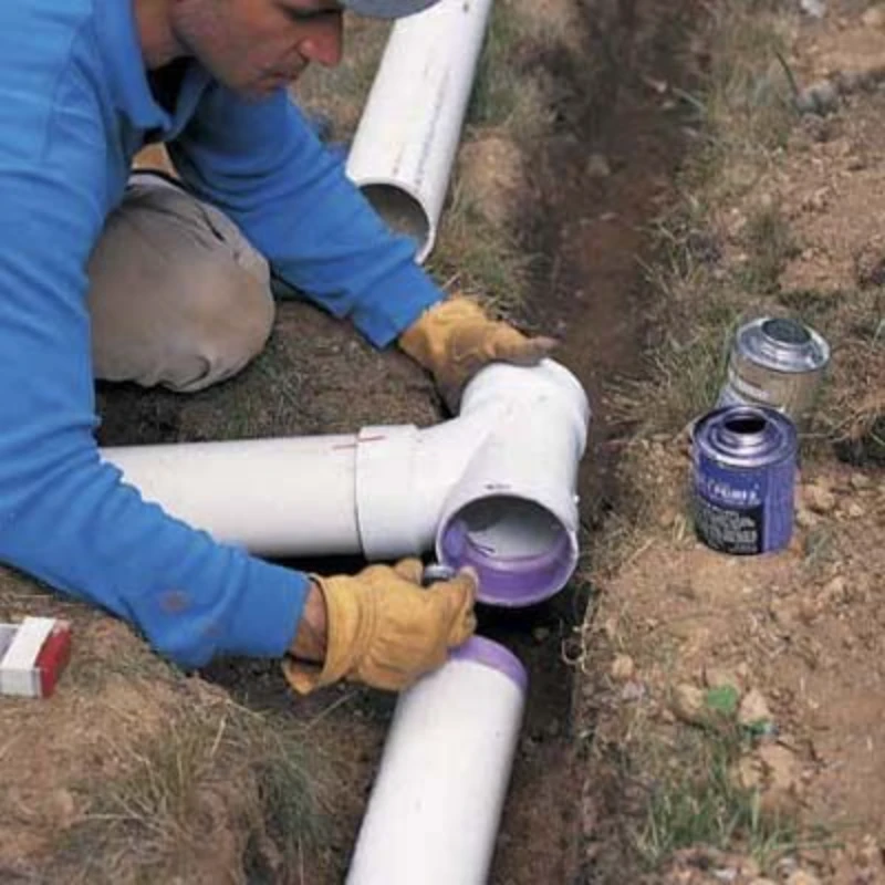 A person in a blue shirt and gloves applies purple PVC primer to connect white pipes in a trench. Nearby are cans of primer and adhesive.