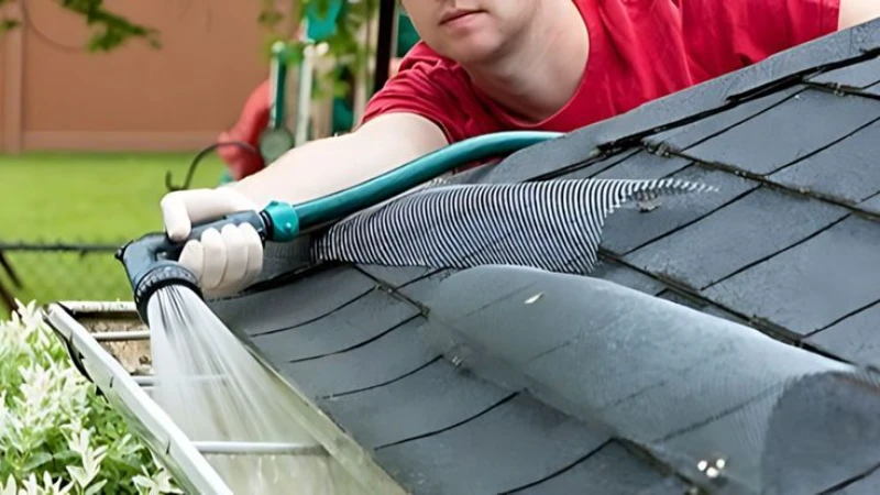 A person in a red shirt uses a hose to clean a house's gray shingled roof. The scene conveys a sense of focus and maintenance effort. Emergency gutter cleaning Dormont, PA
