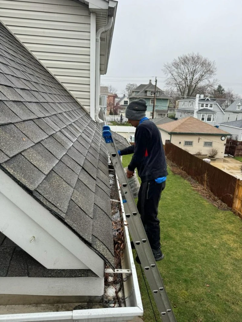 A person stands on a ladder cleaning leaves from house gutters on a gray, overcast day. The scene features suburban homes and a grassy yard. Seamless gutter cleaning Dormont, PA
