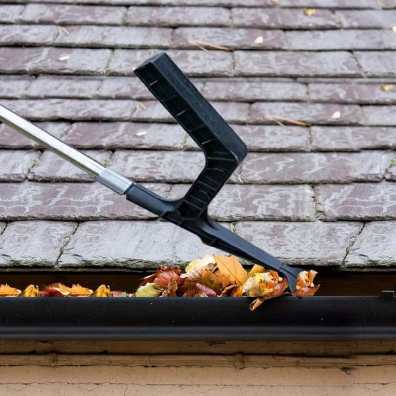 A tool clears autumn leaves from a roof gutter on a shingled rooftop, emphasizing maintenance and seasonal cleaning.