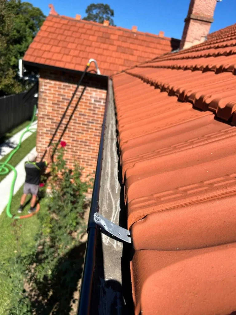 A person uses a long tool to clean gutters on a red-tiled roof under a clear blue sky. The scene conveys industriousness and maintenance.