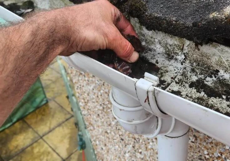 A hand cleaning debris from a white gutter attached to a textured wall. The scene conveys routine maintenance, with dirt and moss visible.