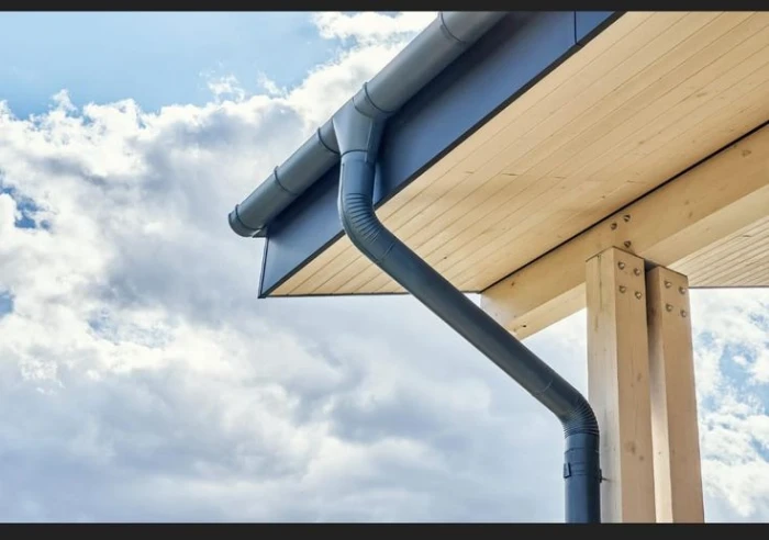 Close-up view of a modern gutter and downspout system attached to the roof of a wooden structure, with a partly cloudy sky in the background. PA