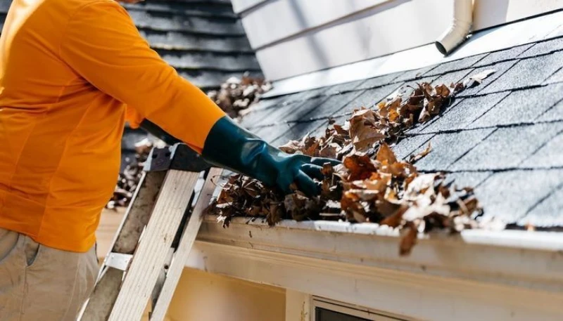 A person wearing a bright orange long-sleeve shirt and dark green protective gloves stands on a ladder to manually scoop dry, brown autumn leaves out of a clogged white roof gutter.