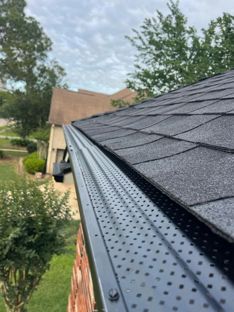 A close-up, high-angle view of a black metal gutter system fitted with a perforated mesh leaf guard along a dark shingled roofline on a brick home. gutter guard installation pa