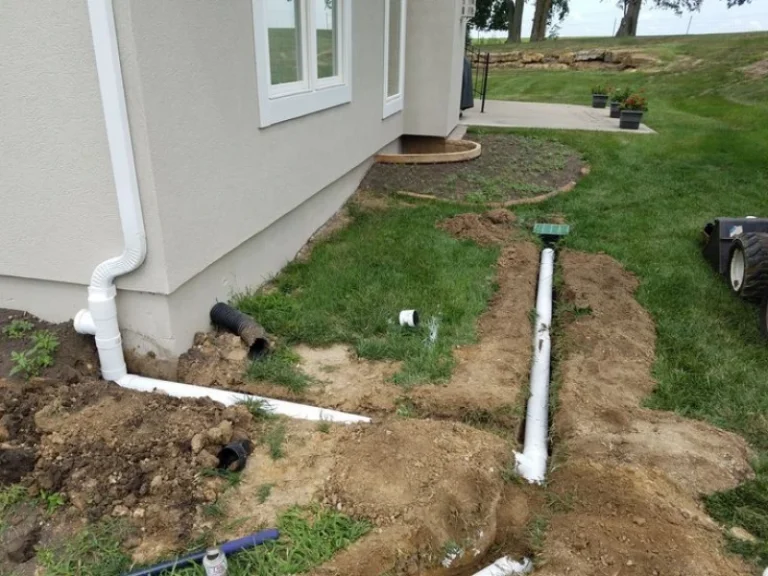 Drainage pipes are being installed around the exterior of a house with a white downspout connected to a trench in the yard. How Much Does It Cost to Install a Downspout?