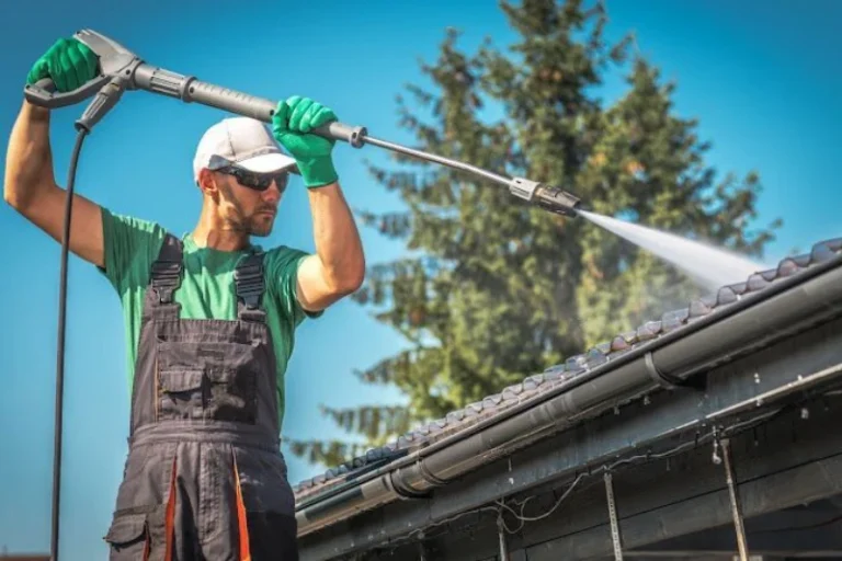 A man cleaning a roof with a pressure washer, wearing sunglasses and gloves. Why You Should Pressure Wash Gutters and Downspouts?