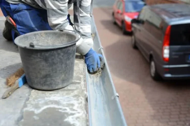 A person wearing blue gloves cleans debris from a residential roof gutter into a black bucket.